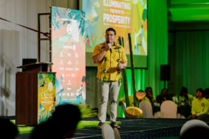 Austin Shelton, Ph.D., wearing a yellow shirt and lei, speaks into a microphone on stage during the 17th UOG Conference on Island Sustainability.