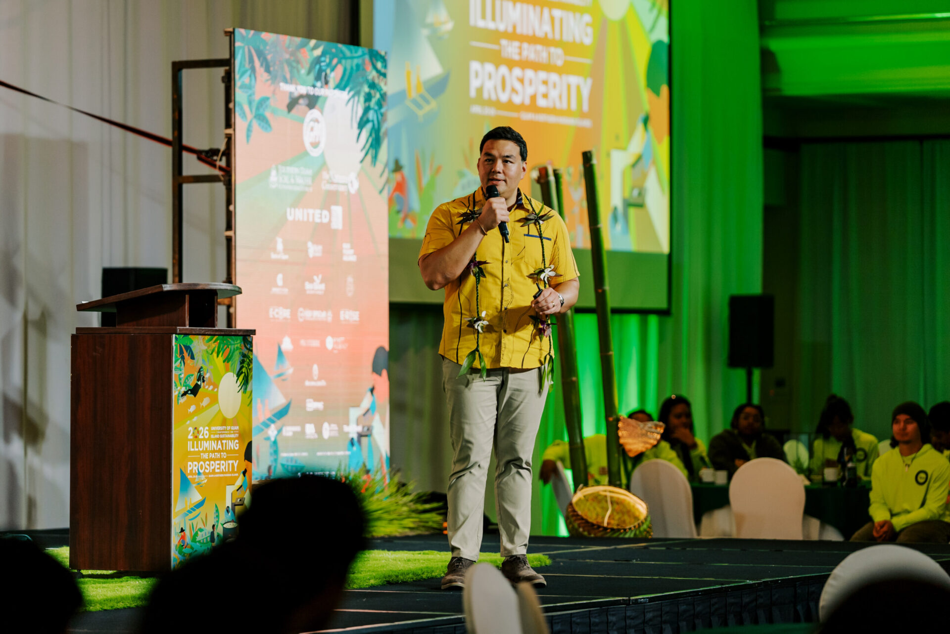 Austin Shelton, Ph.D., wearing a yellow shirt and lei, speaks into a microphone on stage during the 17th UOG Conference on Island Sustainability.