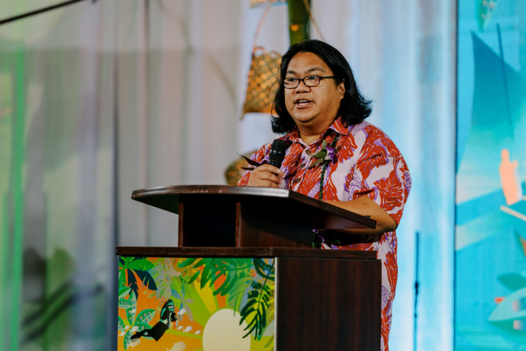 William Castillo, wearing a red floral aloha shirt and a lei, speaks into a microphone behind a tropical-themed podium at the 17th UOG Conference on Island Sustainability.