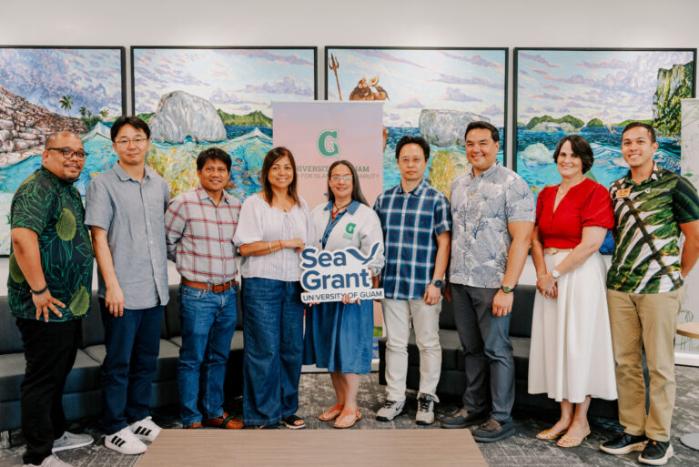 A group of nine University of Guam researchers and leaders standing together and holding a "Sea Grant University of Guam" sign in front of large landscape paintings.