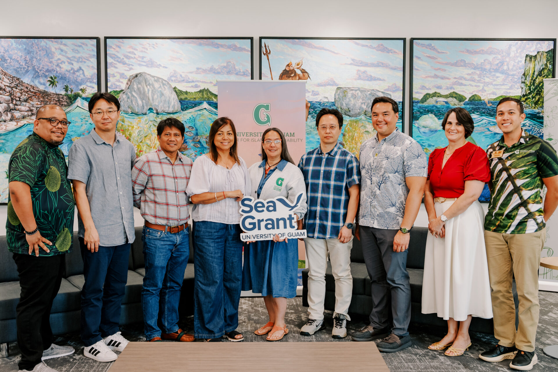 A group of nine University of Guam researchers and leaders standing together and holding a "Sea Grant University of Guam" sign in front of large landscape paintings.