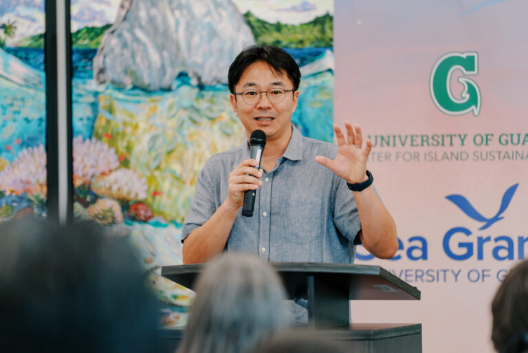 Dr. Chris Yeo gestures while speaking into a microphone at a podium during the UOG Sea Grant research funding announcement.