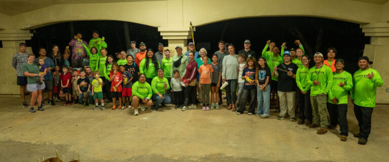 A large group photo of workshop participants and volunteers, many wearing bright green shirts, posing together at night under an open-air pavilion while holding up brown treesnakes captured during an evening hunt.