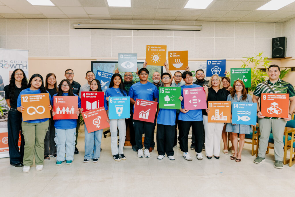 A large group of youth conservation corps members and leadership pose indoors, holding up colorful square signs with various icons and Chamoru text representing local sustainability and conservation goals.