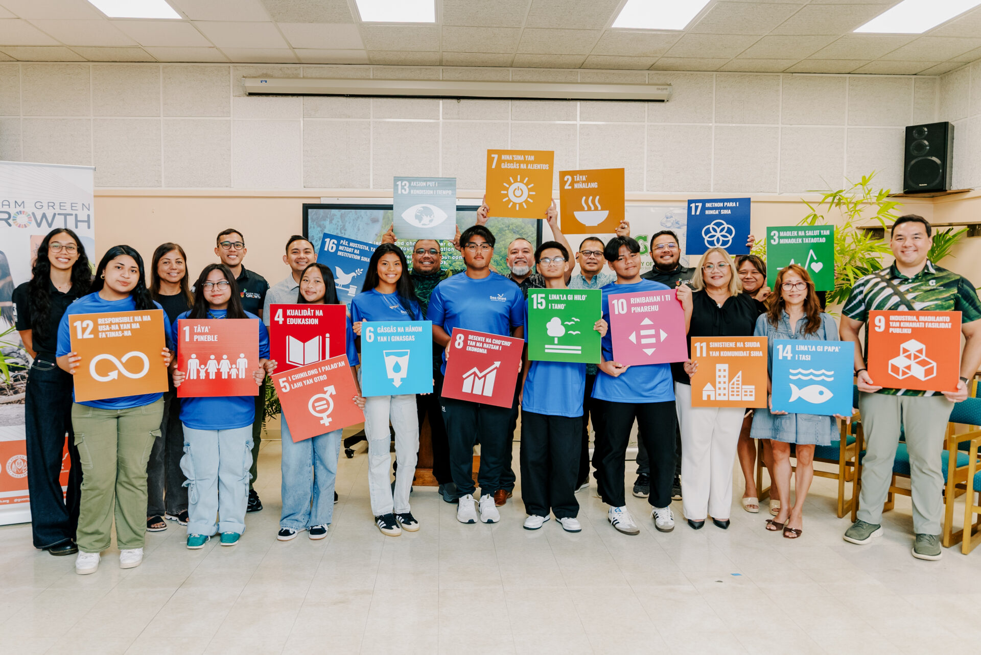 A large group of youth conservation corps members and leadership pose indoors, holding up colorful square signs with various icons and Chamoru text representing local sustainability and conservation goals.