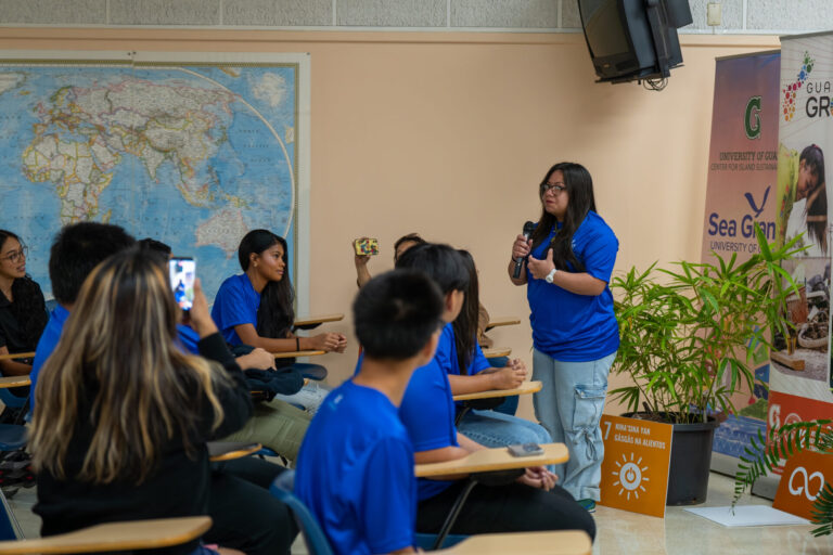 A young female student wearing a blue t-shirt stands and speaks into a microphone during a presentation inside a classroom setting at the UOG RFK Library, while other seated youth conservation corps members listen attentively and record with smartphones.