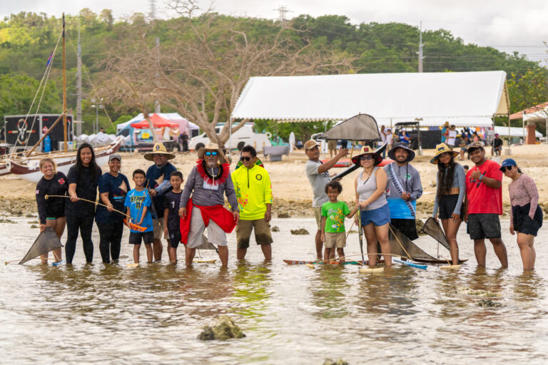 A group poses in the ocean at the Piti Sea Festival mini regatta, with small hand-crafted galaide’ proa models with black sails visible in the shallow water. Spectators and event tents are in the background on the beach.