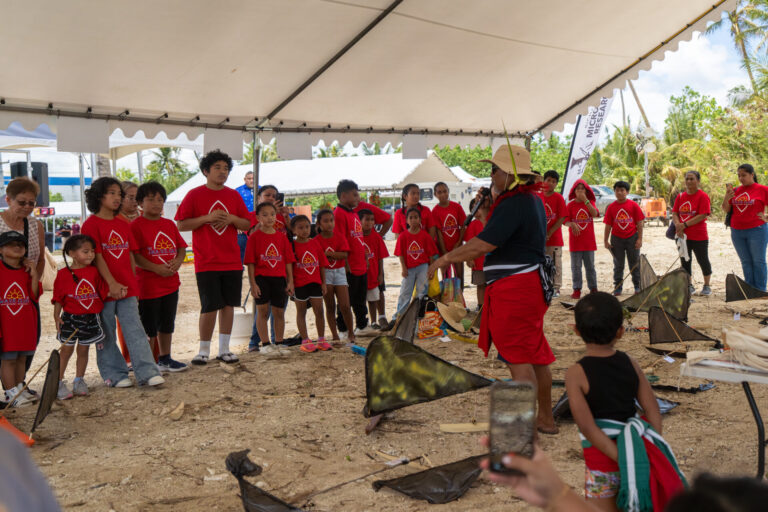 Master Navigator Larry Raigatel addresses a group of students in matching red shirts under a large canopy tent, speaking into a microphone near several small proa models on the ground.