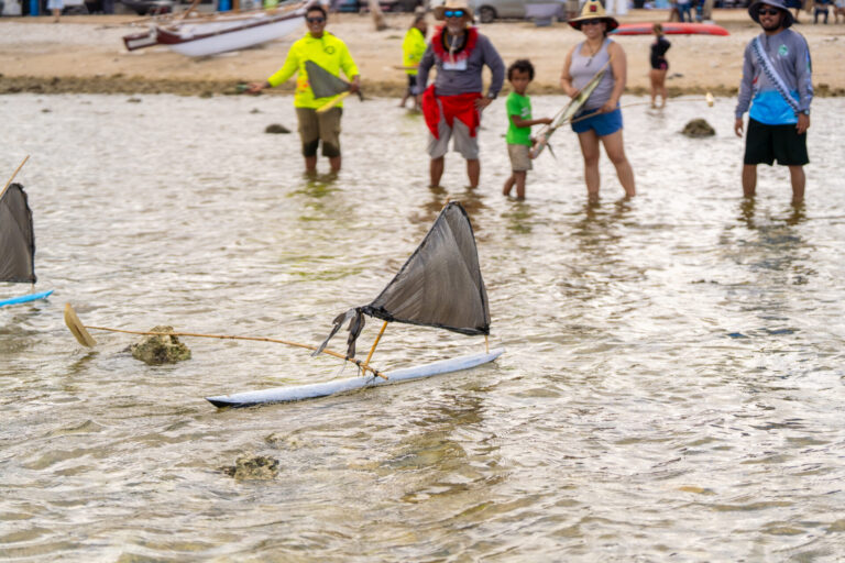 Here is a short alt text for the file "UOG Proa 3.jpg": A small hand-crafted proa model with a black triangular sail floats in the shallow, clear ocean water during a mini regatta competition, with several adult and child spectators watching from the background.