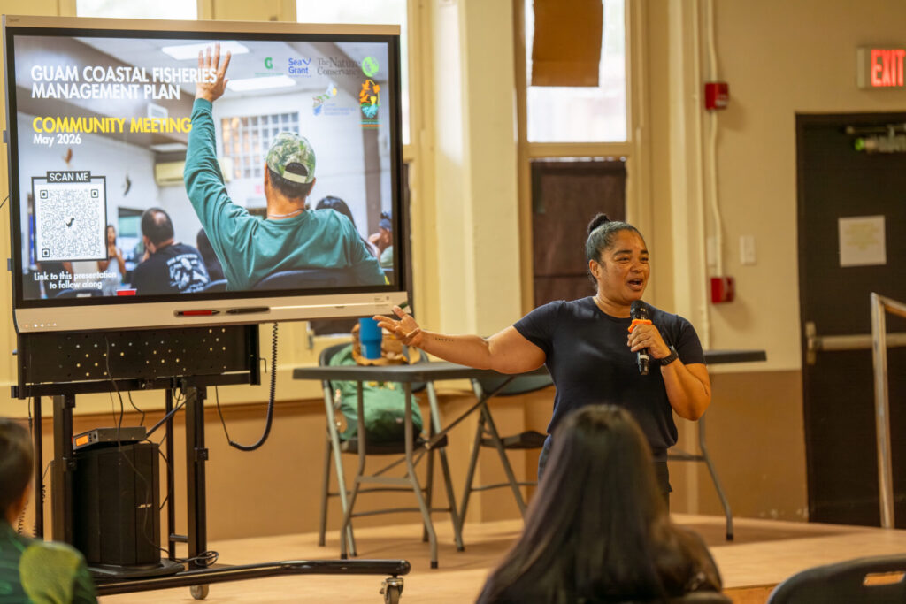 Guam Department of Agriculture Director Chelsa Muna stands indoors speaking into a microphone, gesturing toward a large digital display screen that reads "Guam Coastal Fisheries Management Plan Community Meeting" next to an audience member.