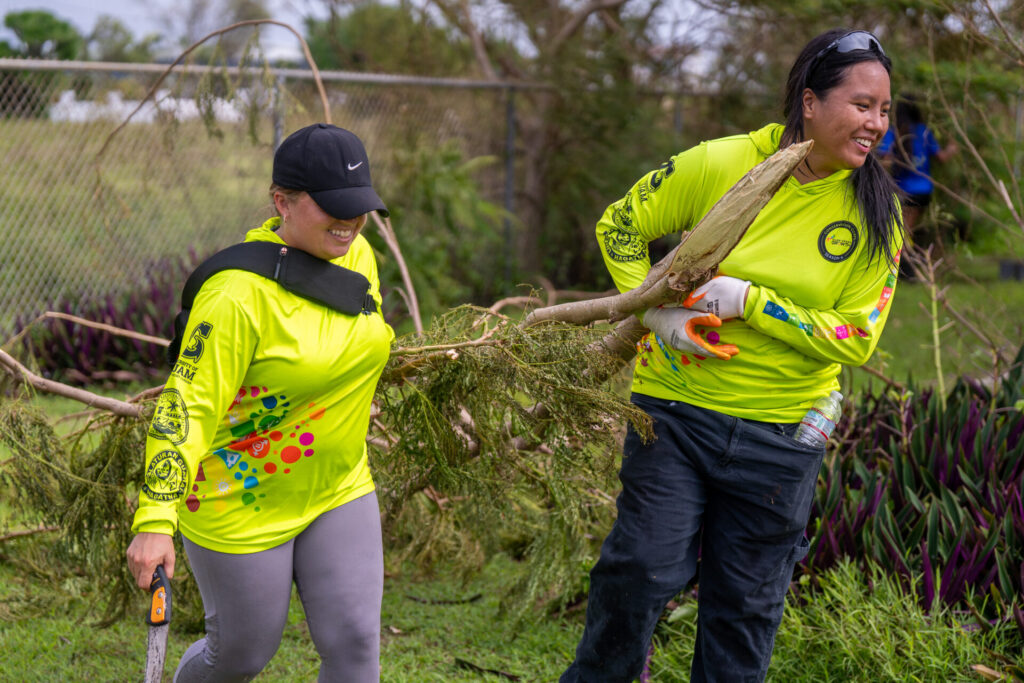 Two members of the Guam Green Growth Conservation Corps, wearing bright neon-yellow long-sleeve shirts, smile as they work together to carry a large, leafy tree branch during cleanup efforts at the UOG Mangilao campus.