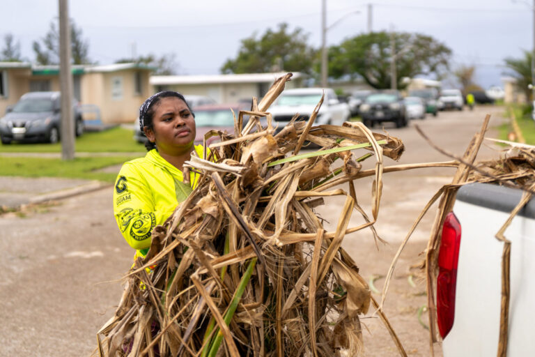 A woman wearing a bright neon-yellow long-sleeve shirt carries a large, heavy bundle of dried plant debris toward the back of a white pickup truck during recovery efforts on a street lined with parked cars.