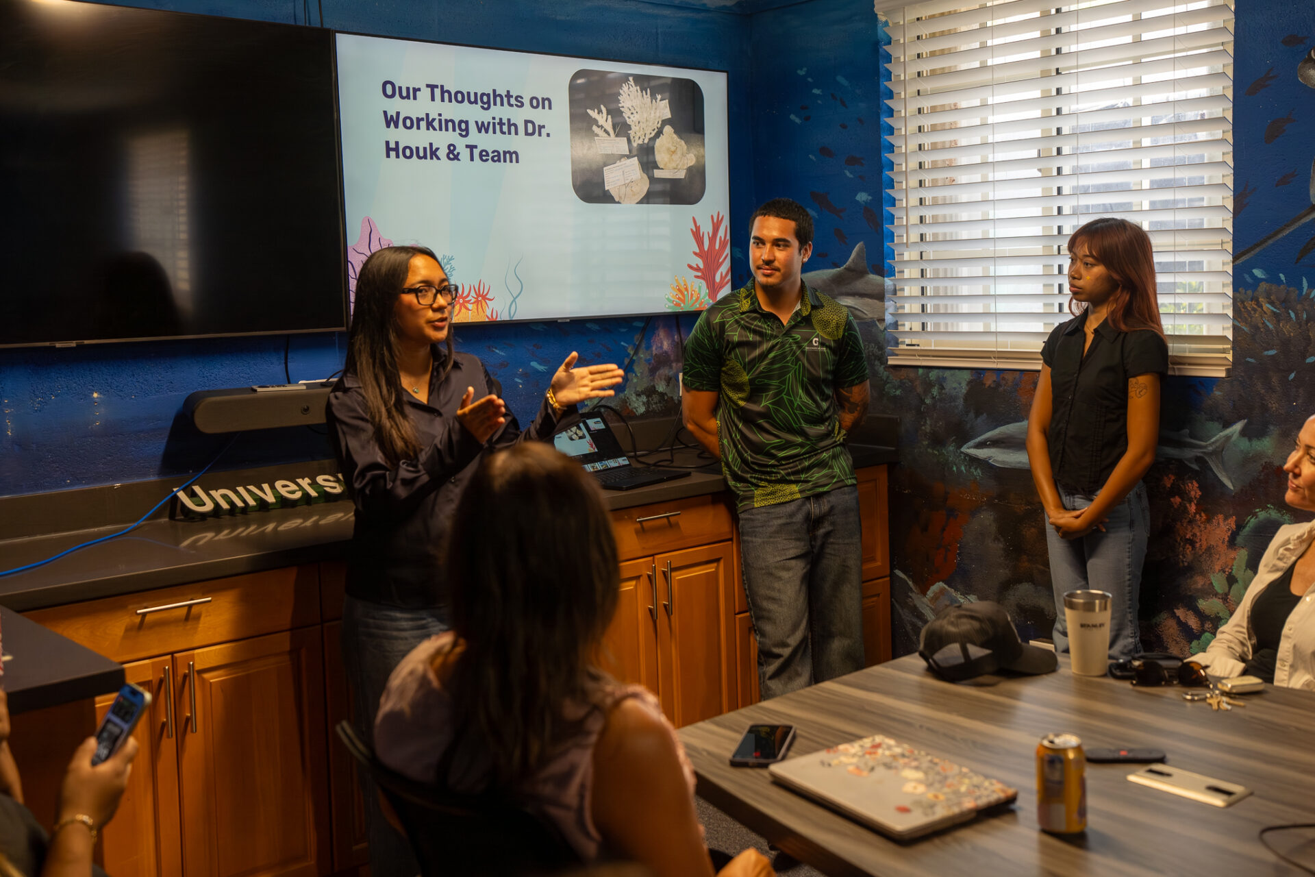 Three student fellows give a presentation in a room with ocean murals. One fellow speaks and gestures in front of a monitor displaying a slide about their coral research titled "Our Thoughts on Working with Dr. Houk & Team," while audience members listen in the foreground.