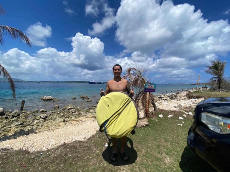 Dominic Torres smiles and gives a shaka sign while standing on a grassy shoreline holding a large, round yellow bag. Behind him is a rocky beach, clear blue ocean, and a bright sky filled with fluffy white clouds.