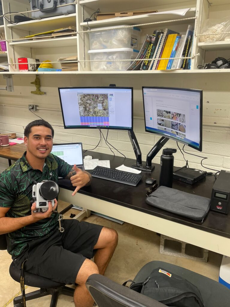 A smiling fellow sits at a lab desk, holding an underwater camera housing and giving a shaka sign. Behind him, two large computer monitors display coral reef imagery and the CoralNet platform used for research data analysis.