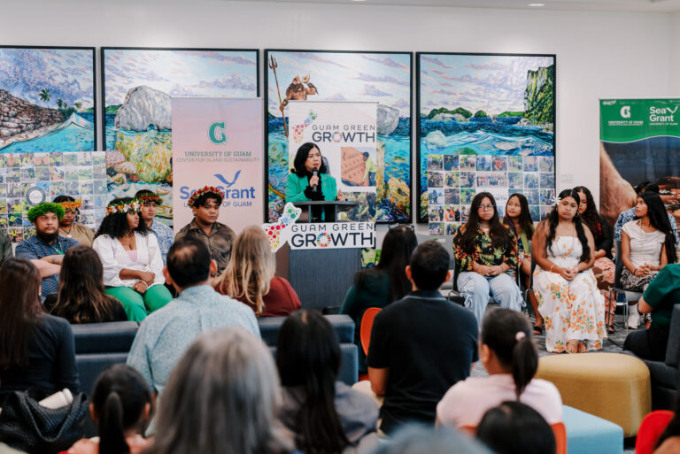 UOG President Anita Borja Enriquez speaking from a Guam Green Growth podium, addressing a seated audience of G3 Conservation Corps graduates and guests.