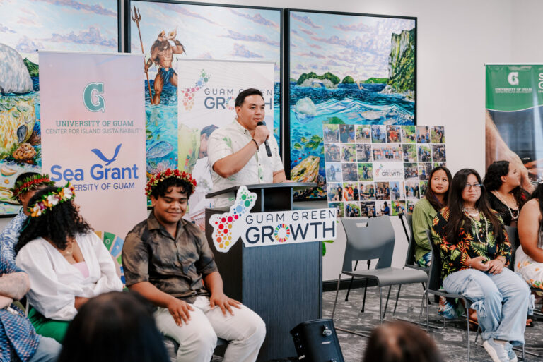 Dr. Austin Shelton speaking at a Guam Green Growth podium, addressing seated G3 Conservation Corps graduates with UOG banners and large island-themed paintings in the background.