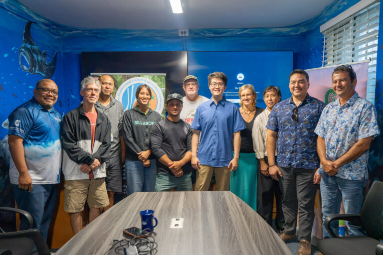 Navigating Home Fellow Enqi Yang stands in the center of a group photo alongside agency mentors and UOG CIS & Sea Grant leadership, including Director Austin Shelton and Associate Director Kyle Mandapat. The group of eleven is gathered behind a conference table in a room featuring a blue, ocean-themed mural.