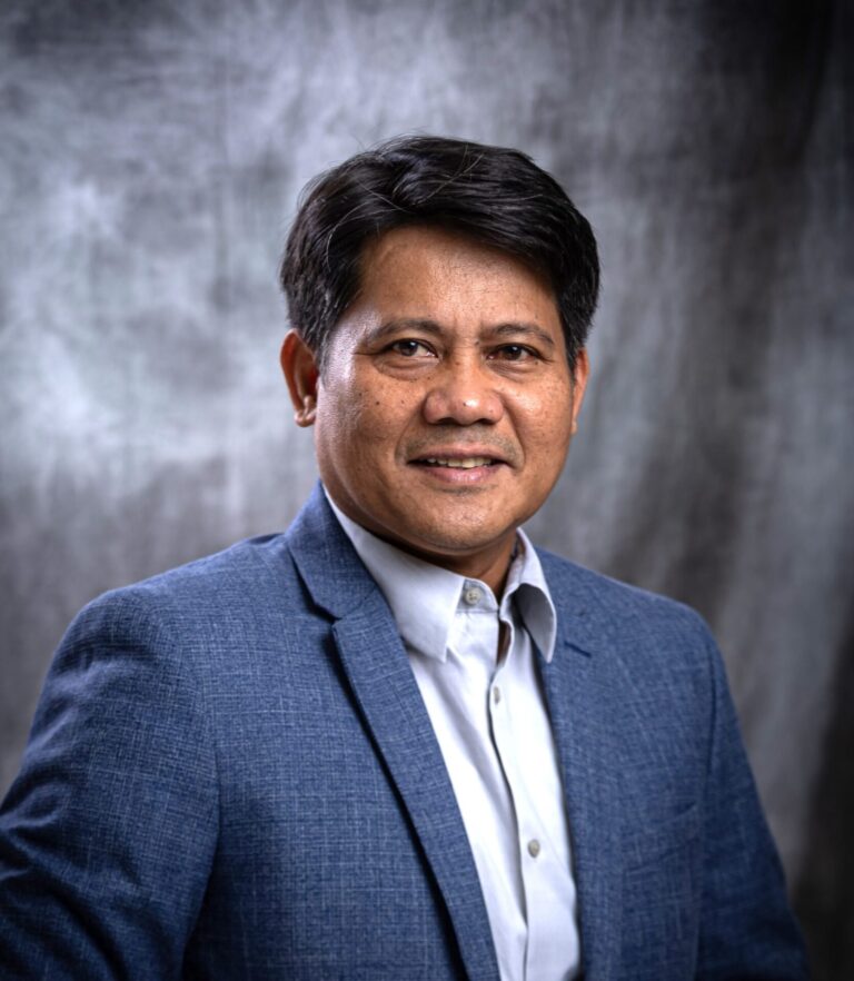 Professional portrait of Ernesto J. Guades, Ph.D., chair and associate professor of civil engineering at the University of Guam, smiling and wearing a blue blazer against a grey textured backdrop.