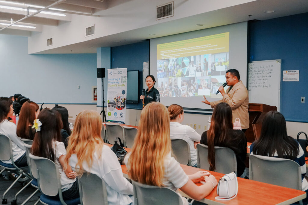G3 Action Framework Coordinators Camarin Quitugua and Jaret Castro hold microphones and speak to a classroom of seated students during a presentation in front of a projection screen.