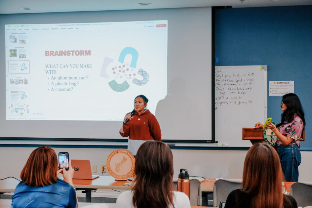A student speaks into a microphone in front of a "BRAINSTORM" presentation slide during a Junior Achievement Guam Masterclass.