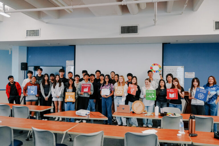 Junior Achievement Guam high school students pose with G3 Makerspace Coordinator Abby Crain while holding colorful UN Sustainable Development Goal posters.
